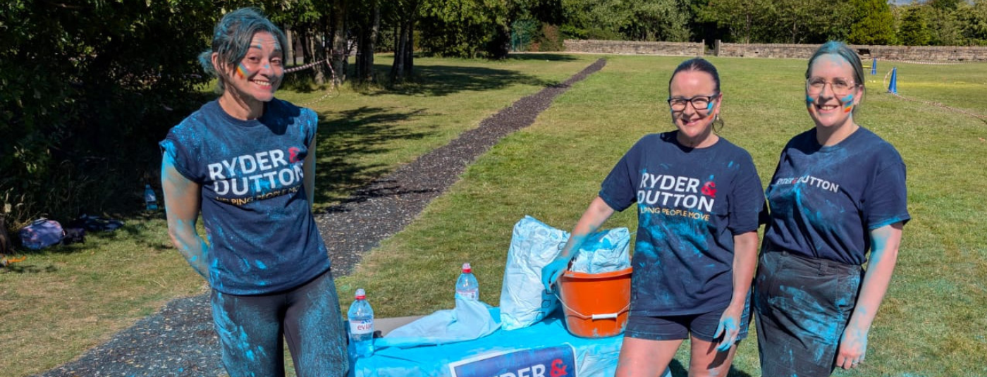 Three fundraisers in matching blue t-shirts with 'Ryder & Dutton' logo at an outdoor colour run event.